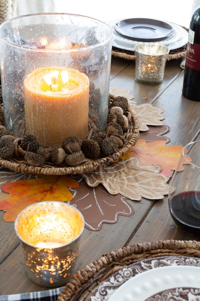 Glass hurricane on a wicker plate charger. Pumpkin color candle and acorns make a nice centerpiece.